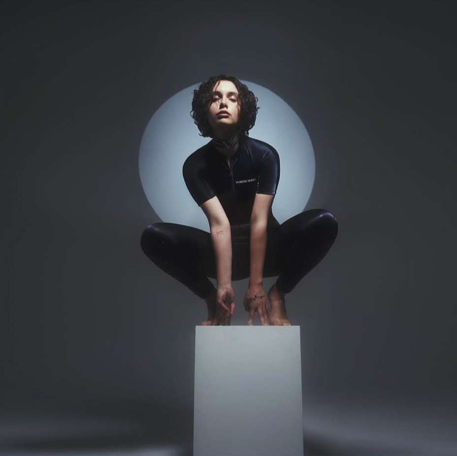 A young adult female with dark hair is crouching on a white cube in a gray studio with a soft round spotlight behind her.