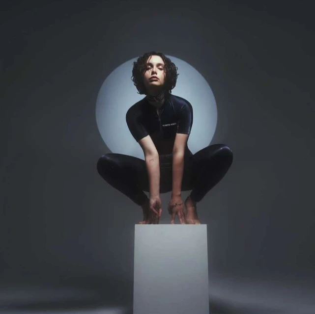 A young adult female with dark hair is crouching on a white cube in a gray studio with a soft round spotlight behind her.
