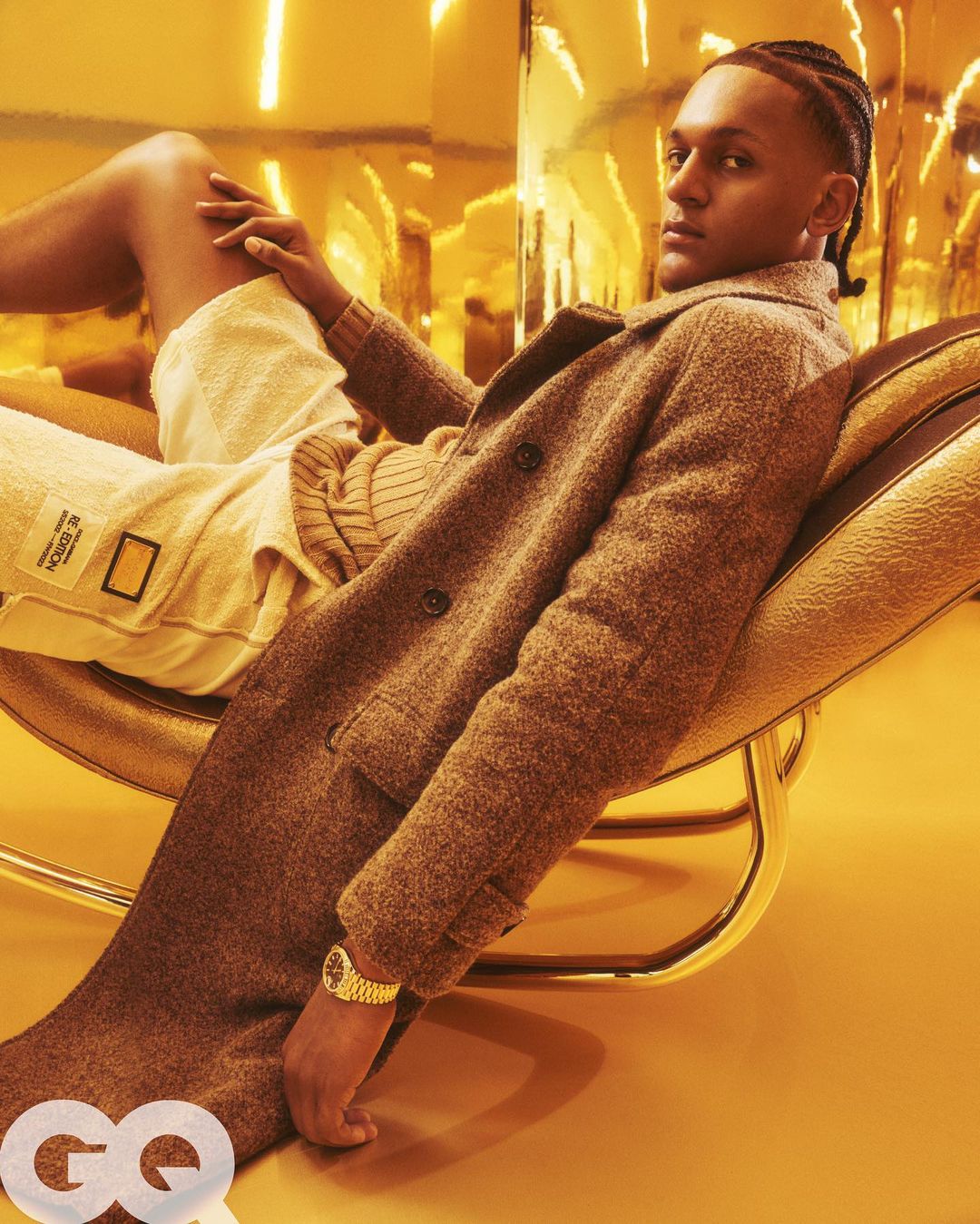 A man lounges on a gold chair wearing a brown coat and shorts, highlighted by a gold-toned background, fashion photography.