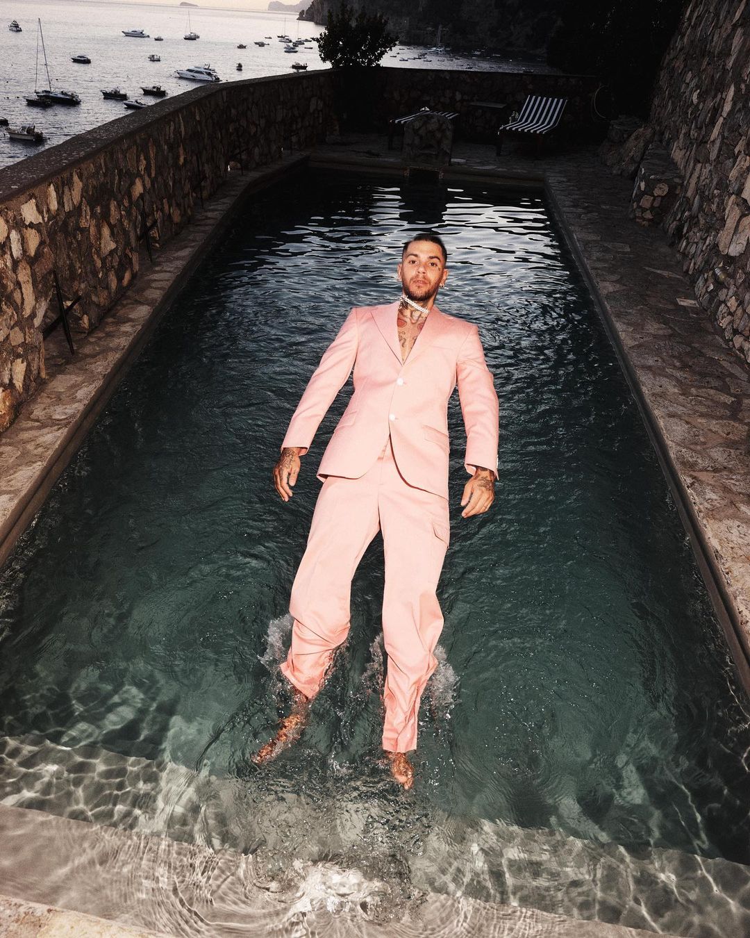 Man in pink suit floats in a pool with boats on the sea in the background under dusky light, creating a surreal and eye-catching scene.