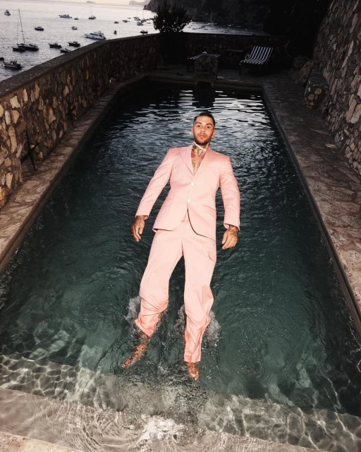 Man in pink suit floats in a pool with boats on the sea in the background under dusky light, creating a surreal and eye-catching scene.