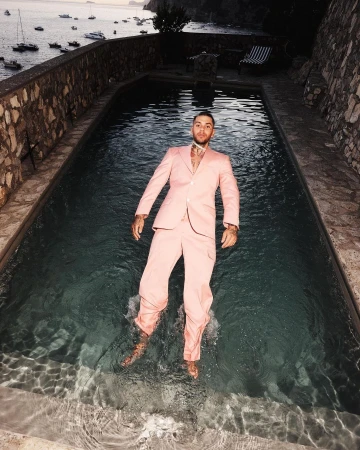 Man in pink suit floats in a pool with boats on the sea in the background under dusky light, creating a surreal and eye-catching scene.