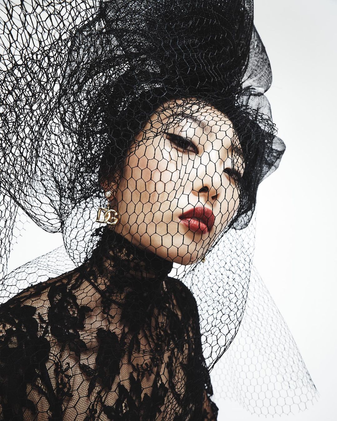 Portrait of an Asian woman wearing a black lace top and large black veil looking directly at the camera in a light studio setting.