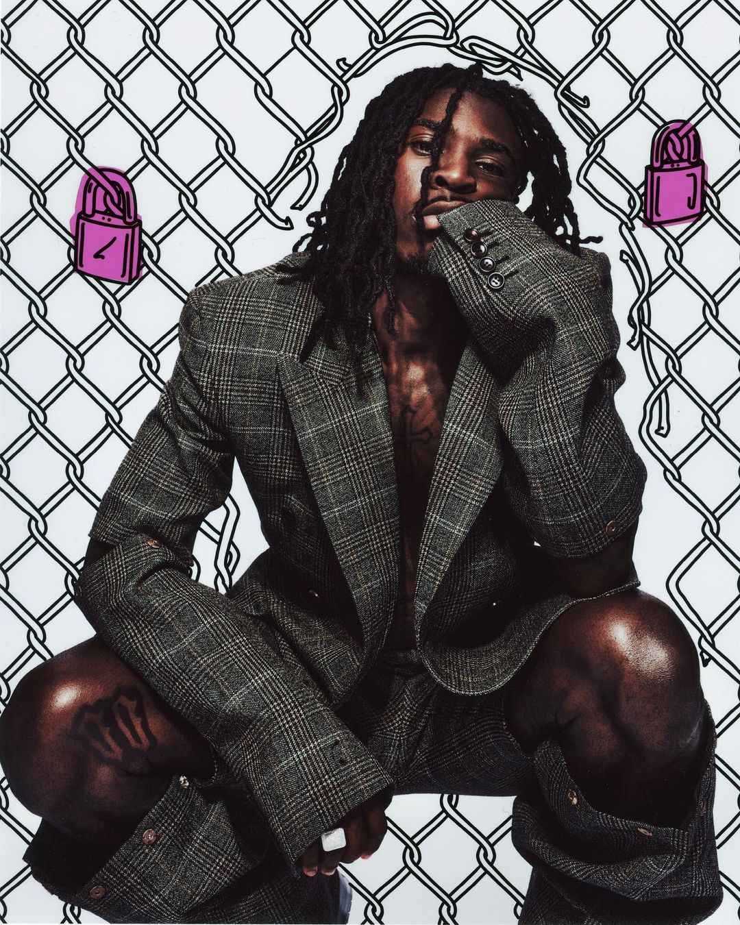 Adult male poses in a grey suit against a wire fence background with pink lock illustrations, creating an edgy fashion statement.