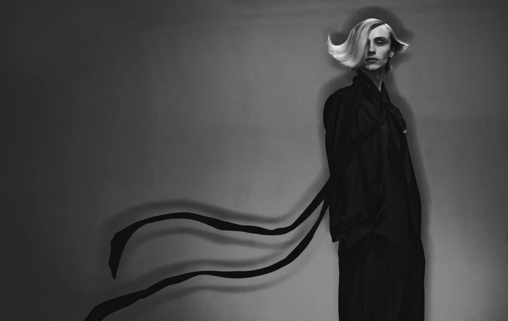 Monochrome fashion portrait of a tall, slender person with a blonde hairstyle, wearing a dark robe and pants against a gray backdrop.
