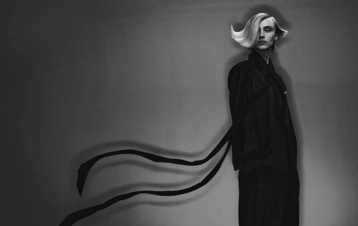 Monochrome fashion portrait of a tall, slender person with a blonde hairstyle, wearing a dark robe and pants against a gray backdrop.