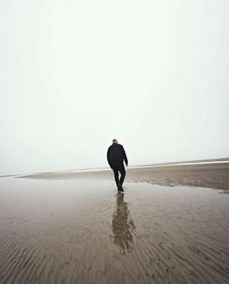 Adult male walks on a wet sandy beach on a foggy day, wearing dark jacket and pants, creating a minimalist image.