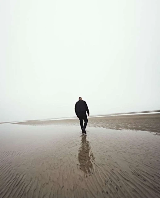 Adult male walks on a wet sandy beach on a foggy day, wearing dark jacket and pants, creating a minimalist image.