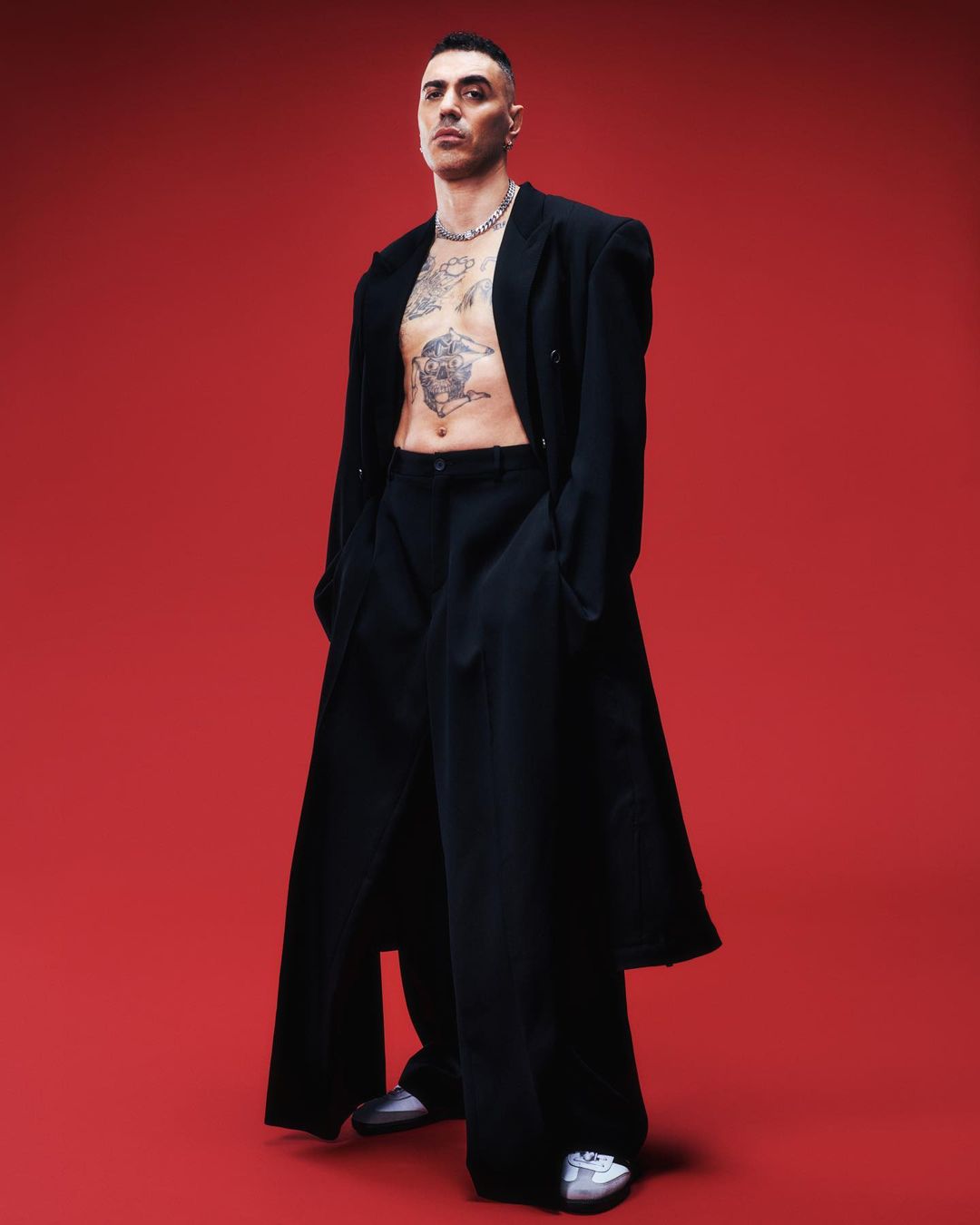 A full-length shot captures a tattooed man in a black coat and pants against a vibrant red background, fashionably posing for the camera.