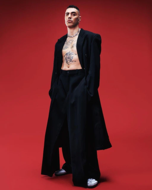 A full-length shot captures a tattooed man in a black coat and pants against a vibrant red background, fashionably posing for the camera.