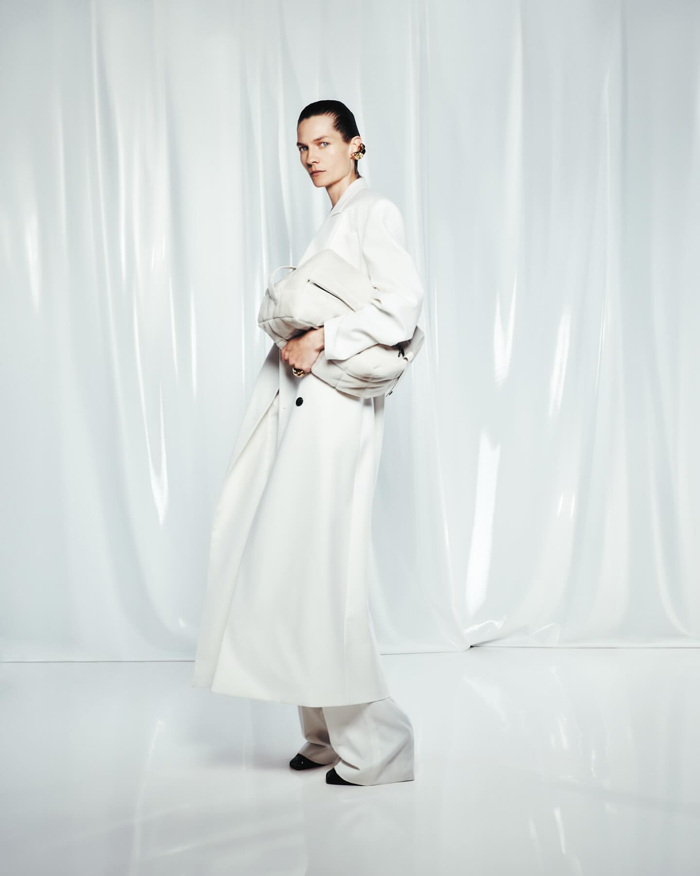 Fashion editorial shot of a woman in a white coat and wide-leg pants holding a clutch, posed in front of a translucent white background.