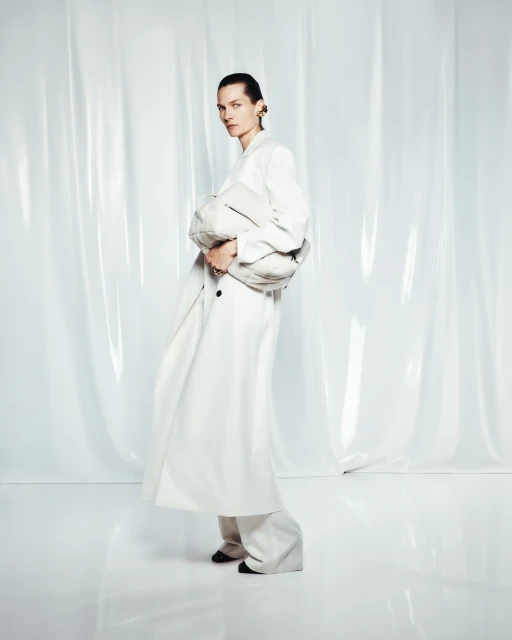 Fashion editorial shot of a woman in a white coat and wide-leg pants holding a clutch, posed in front of a translucent white background.