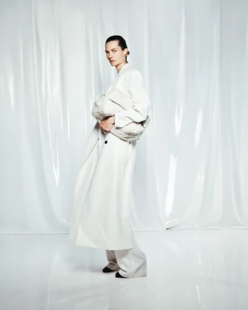 Fashion editorial shot of a woman in a white coat and wide-leg pants holding a clutch, posed in front of a translucent white background.