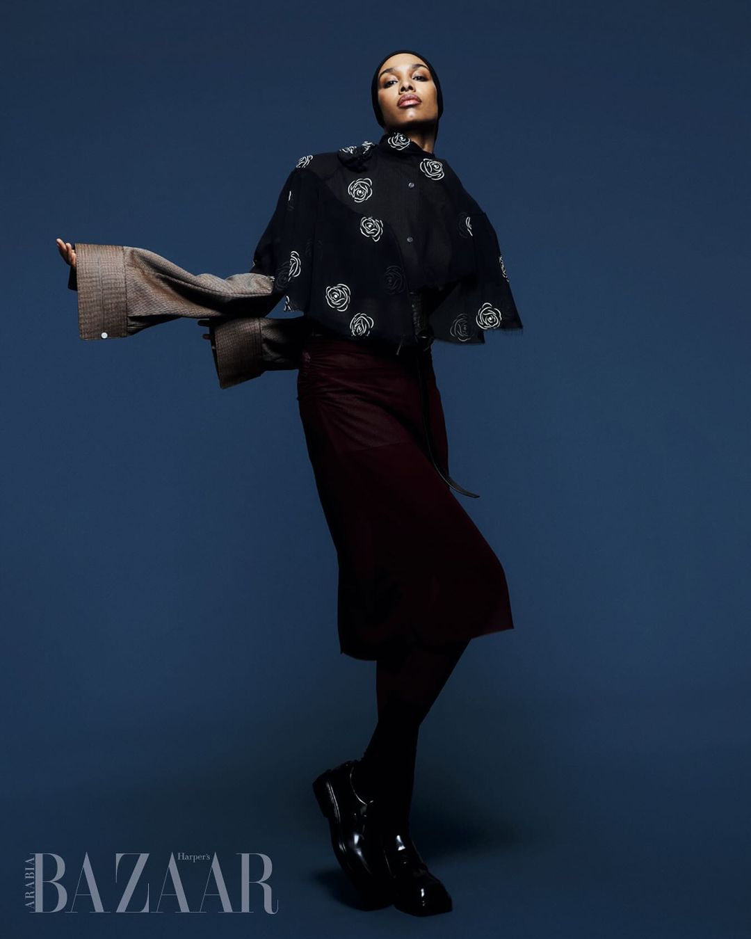 A stylish portrait features a woman in a black floral top and maroon skirt against a blue backdrop, showcasing a high-fashion pose and attire.