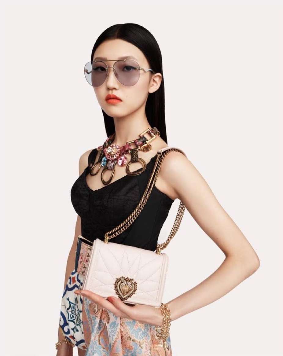 Elegant Asian woman poses in studio with colorful statement necklace, quilted shoulder bag, aviator sunglasses, and patterned skirt.