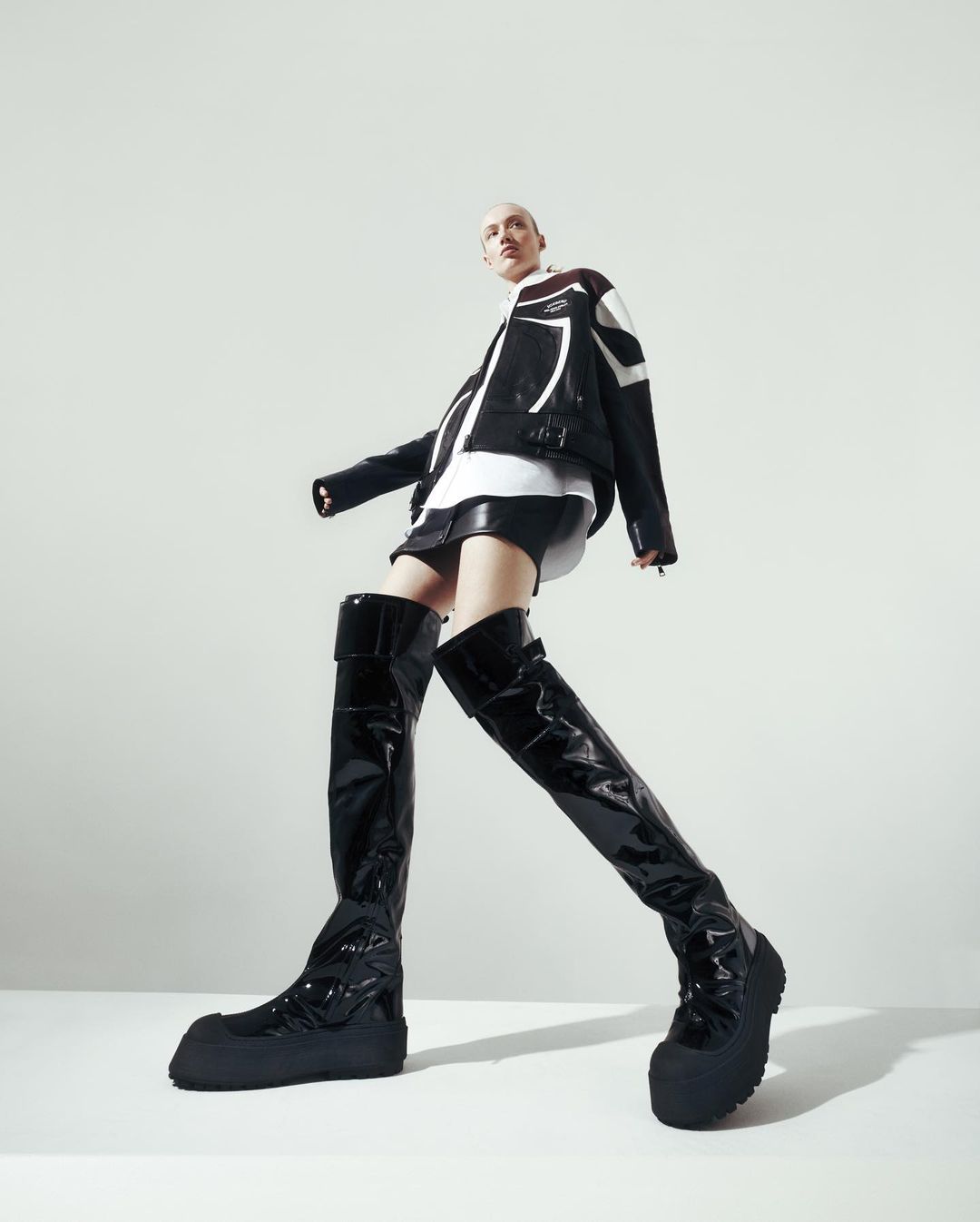A person with shaved head wearing a black and white jacket, black shorts, and shiny black thigh-high boots standing against a plain white background.