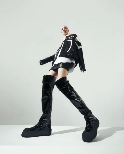 A person with shaved head wearing a black and white jacket, black shorts, and shiny black thigh-high boots standing against a plain white background.