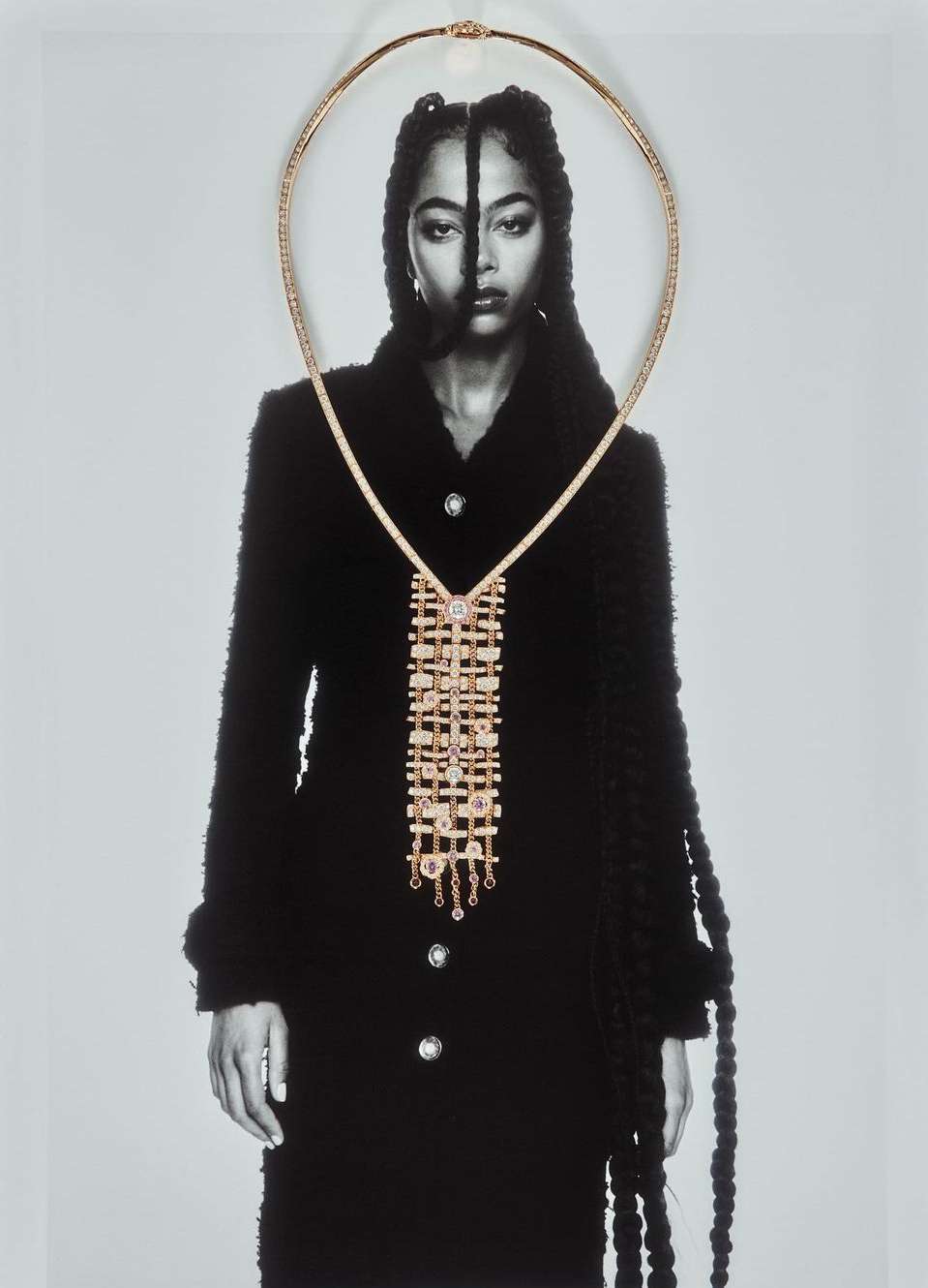 A monochrome photograph of a model wearing a black coat and gold jewelry with a necklace draped over the face and coat.
