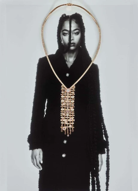 A monochrome photograph of a model wearing a black coat and gold jewelry with a necklace draped over the face and coat.