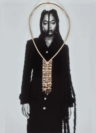 A monochrome photograph of a model wearing a black coat and gold jewelry with a necklace draped over the face and coat.