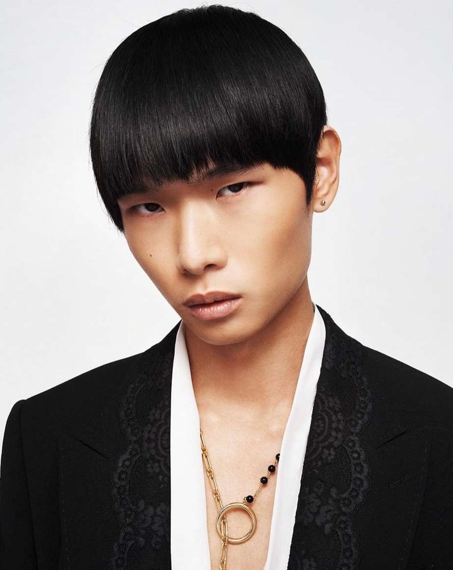 Close-up portrait of a young adult with black hair and a black suit against a white backdrop, focusing on fashion and appearance.