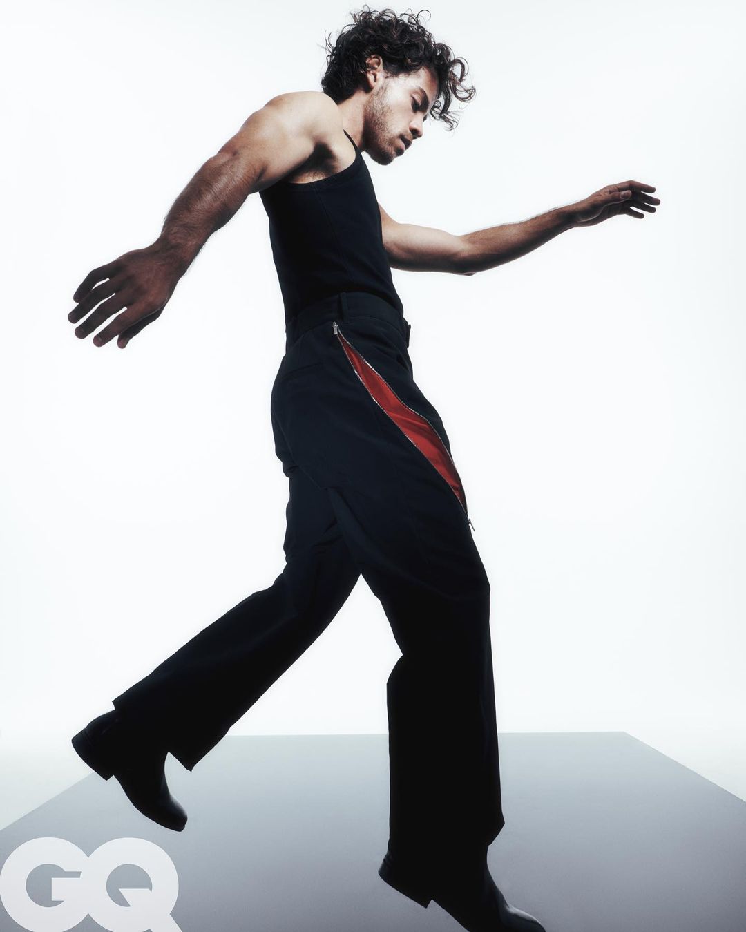 Man strikes a walking pose in a black tank top and pants with a red stripe against a bright white background in a fashion studio setting.