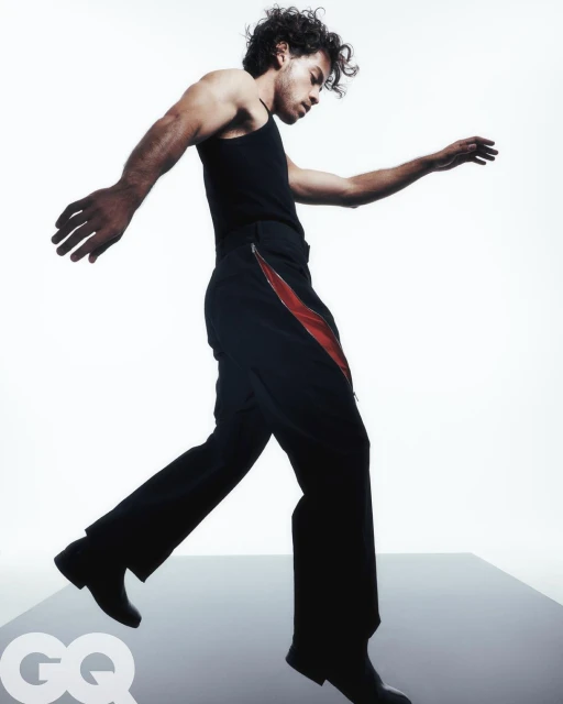 Man strikes a walking pose in a black tank top and pants with a red stripe against a bright white background in a fashion studio setting.
