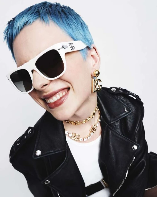 Close-up of a smiling woman with blue hair wearing sunglasses, a black leather jacket, and jewelry against a white background.
