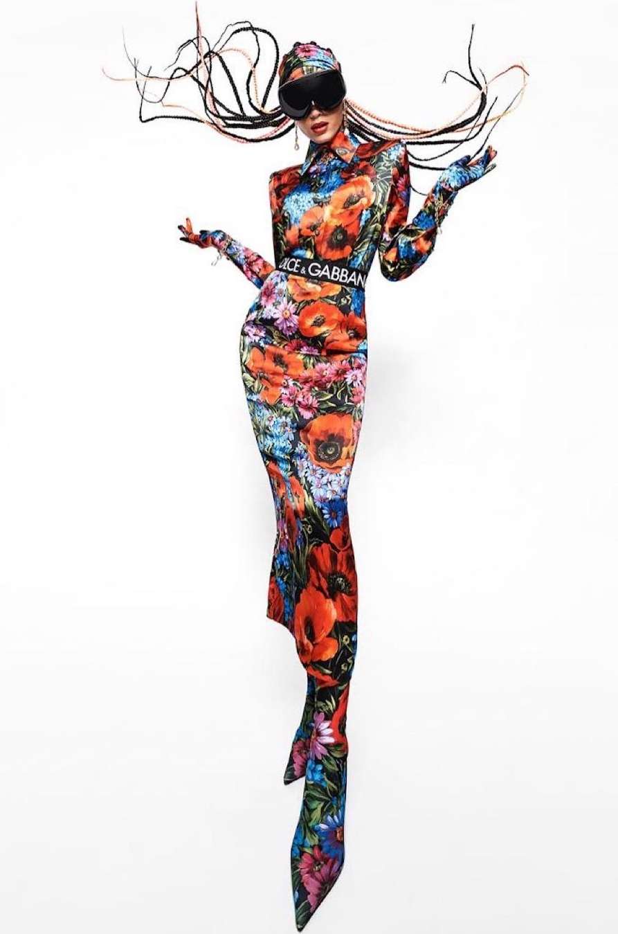 Fashion model wears a full-length floral dress, sunglasses, and gloves, posing in front of a white backdrop with a unique, artistic flair.