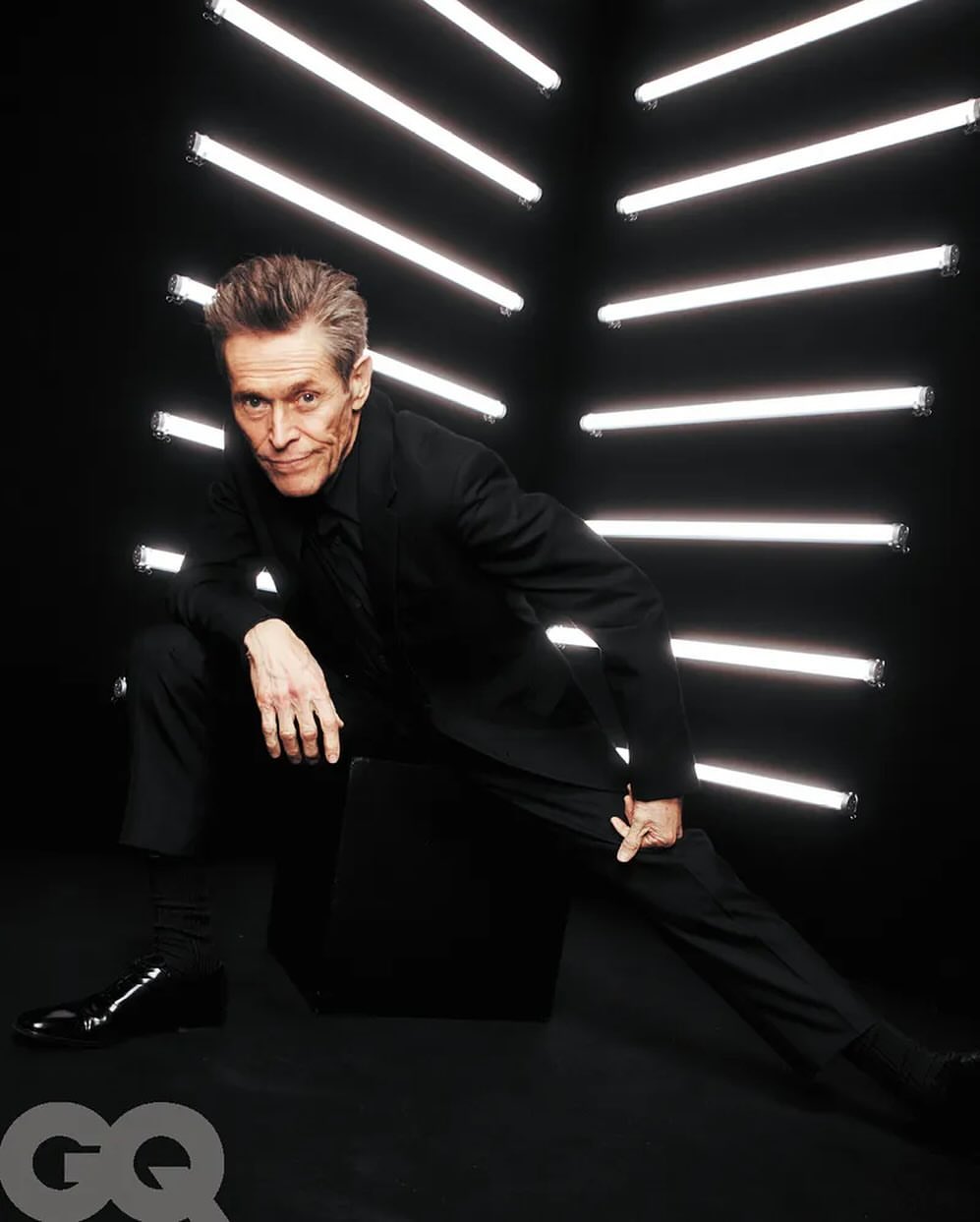 Willem Dafoe in a black suit strikes a dynamic pose in front of geometric neon lights, GQ logo visible, evoking celebrity and entertainment.