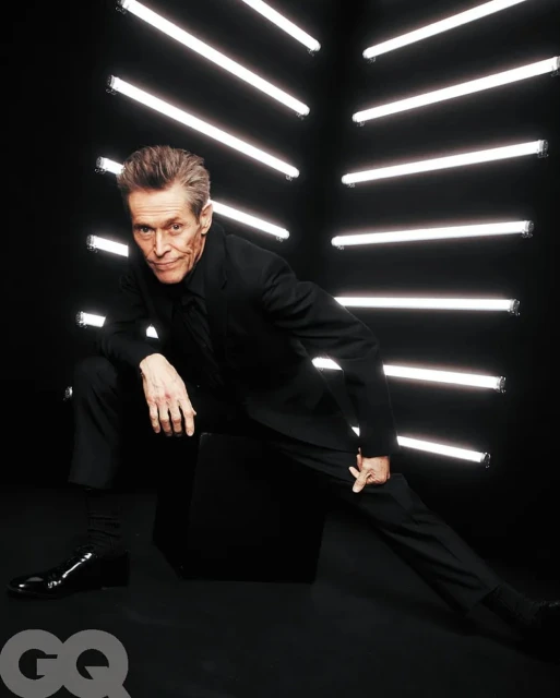 Willem Dafoe in a black suit strikes a dynamic pose in front of geometric neon lights, GQ logo visible, evoking celebrity and entertainment.