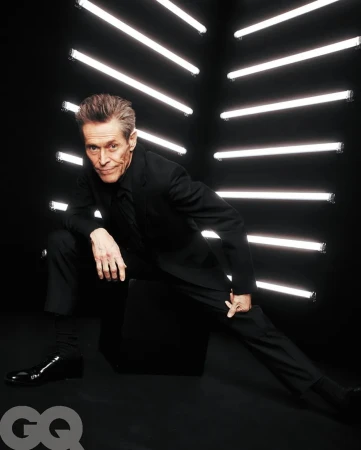 Willem Dafoe in a black suit strikes a dynamic pose in front of geometric neon lights, GQ logo visible, evoking celebrity and entertainment.