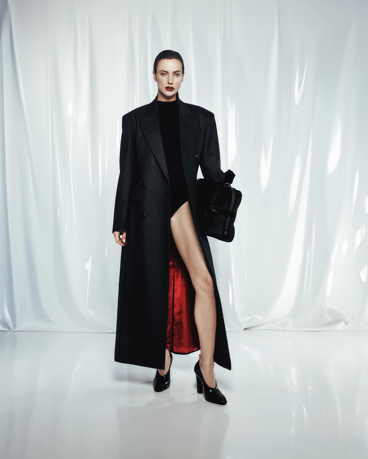 Fashion portrait of an adult female in a black long coat and black pumps, holding a black bag, against a white backdrop.