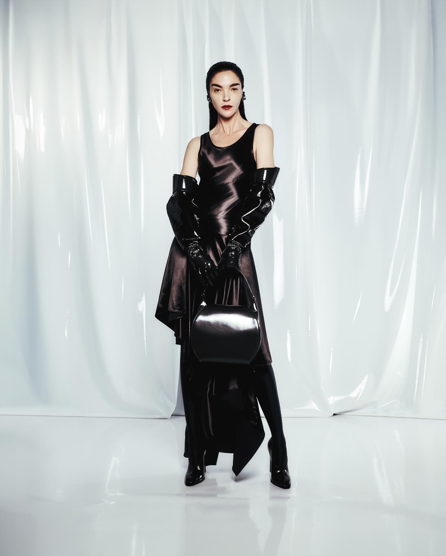 A slender woman in a black satin dress and long gloves poses with a matching purse against a glossy white backdrop in a fashion shot.