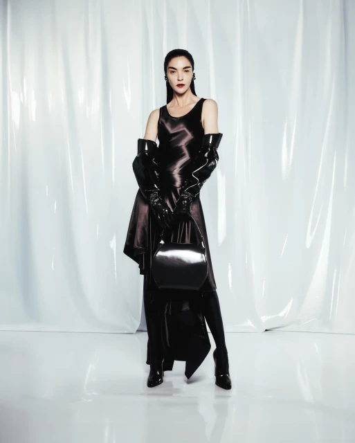A slender woman in a black satin dress and long gloves poses with a matching purse against a glossy white backdrop in a fashion shot.