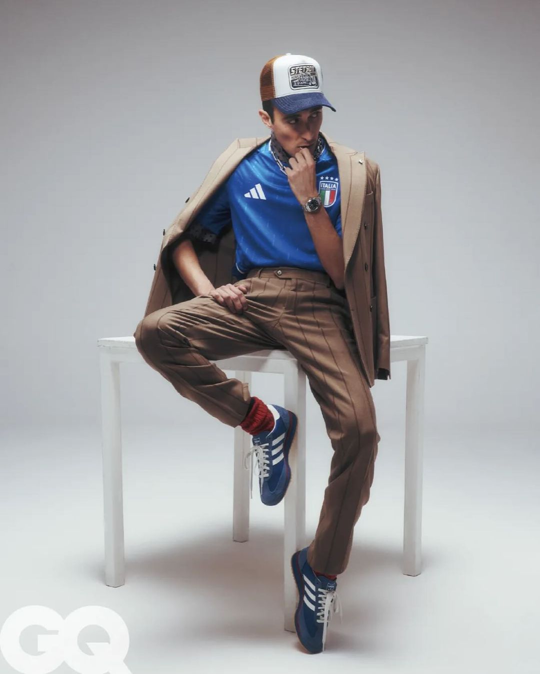 Man seated on a white table wearing a blue jersey, brown trousers, blue sneakers, and a trucker hat with a jacket draped over his shoulders.
