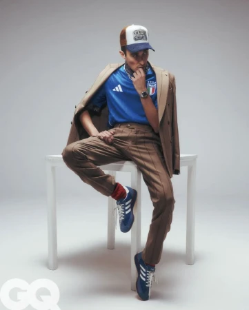 Man seated on a white table wearing a blue jersey, brown trousers, blue sneakers, and a trucker hat with a jacket draped over his shoulders.