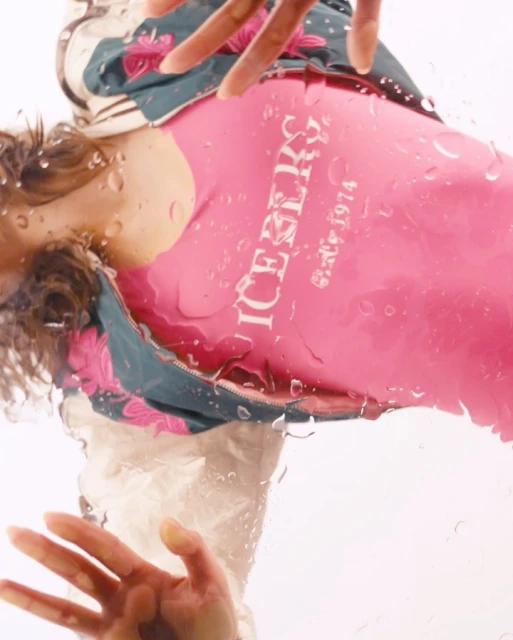 Model in a pink top poses behind water droplets on glass, showcasing a floral jacket and hands with a bright, clean aesthetic.