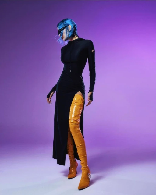 Stylish woman with blue hair poses in a black dress and yellow thigh-high boots against a graduated purple backdrop in a studio setting.
