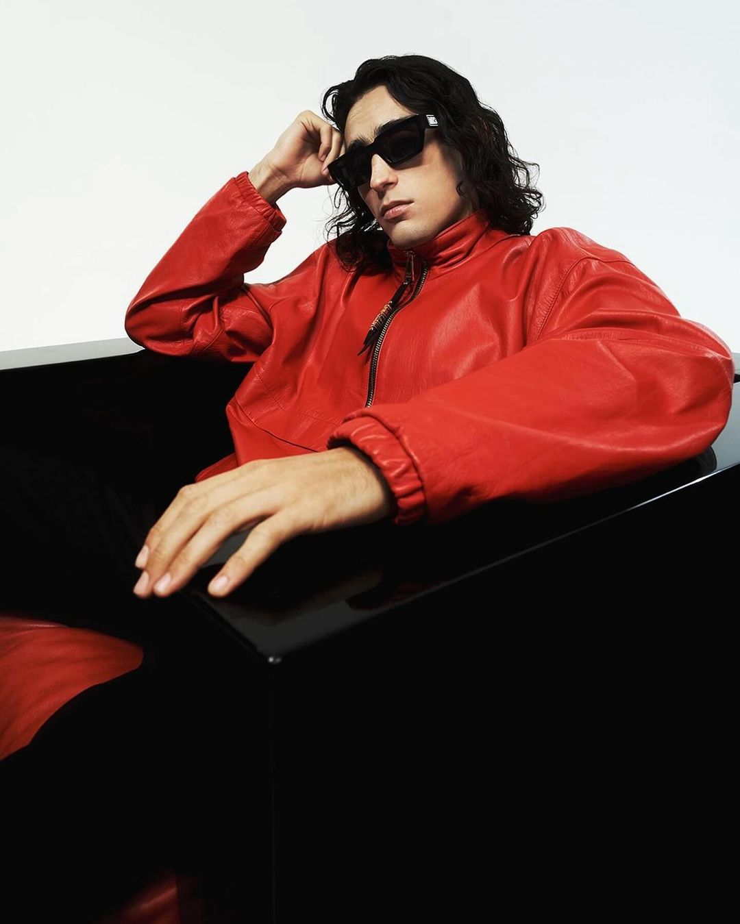 Man with long dark hair poses in sunglasses and a red leather jacket sitting against a glossy black surface in a studio shot.