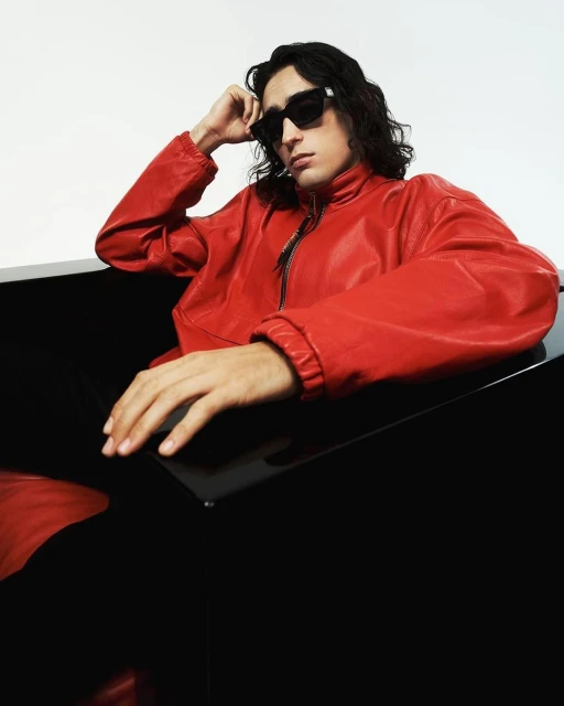 Man with long dark hair poses in sunglasses and a red leather jacket sitting against a glossy black surface in a studio shot.