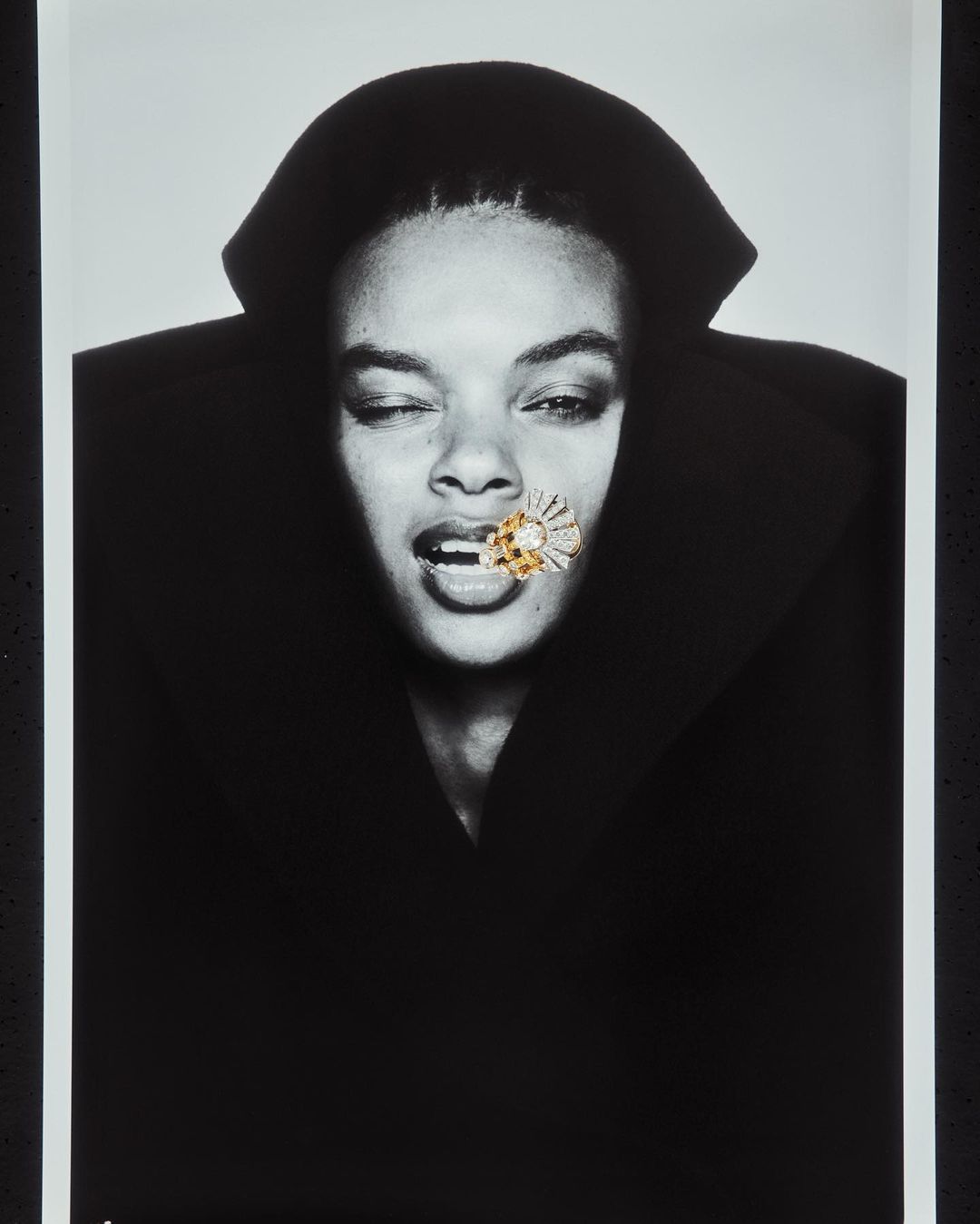 Black and white portrait of a woman winking with a diamond flower embellishment in her mouth against a bright backdrop.