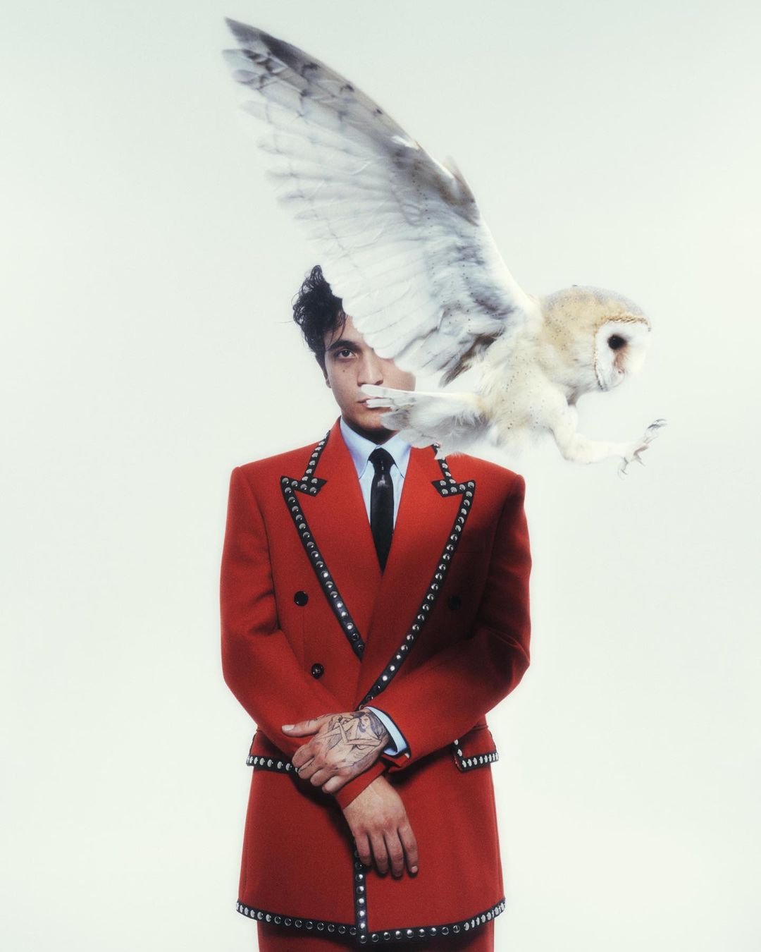 Man in red suit with studded trim poses with a white owl in a portrait, set against a bright white background. Fashion and wildlife meet.