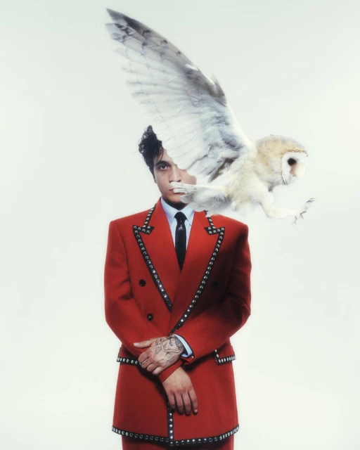 Man in red suit with studded trim poses with a white owl in a portrait, set against a bright white background. Fashion and wildlife meet.