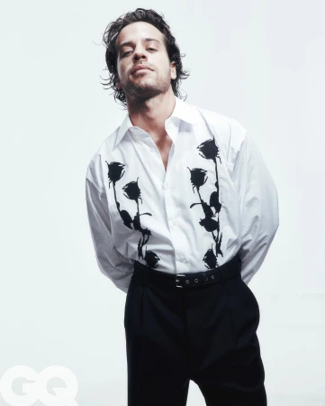 Adult man in a white button-down shirt with a rose print and black pants stands indoors against a white backdrop in a studio shot.