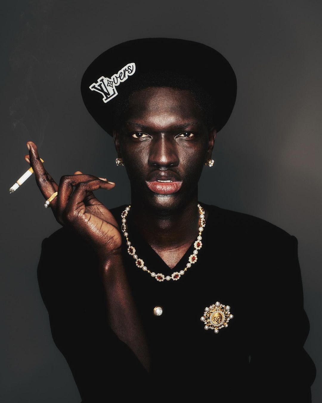 Portrait of a dark-skinned adult male in a black suit and hat smoking a cigarette while wearing jeweled necklace, earrings, and brooch.