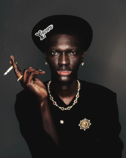 Portrait of a dark-skinned adult male in a black suit and hat smoking a cigarette while wearing jeweled necklace, earrings, and brooch.
