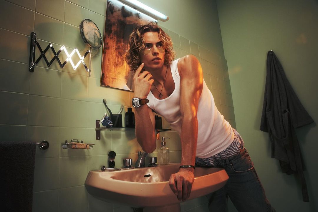 An adult male with long blond hair leans against a sink in a bathroom with green tiled walls and a vintage aesthetic. 