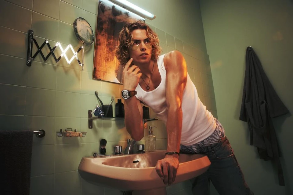 An adult male with long blond hair leans against a sink in a bathroom with green tiled walls and a vintage aesthetic. 