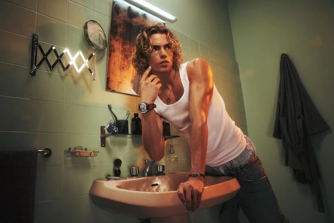 An adult male with long blond hair leans against a sink in a bathroom with green tiled walls and a vintage aesthetic. 